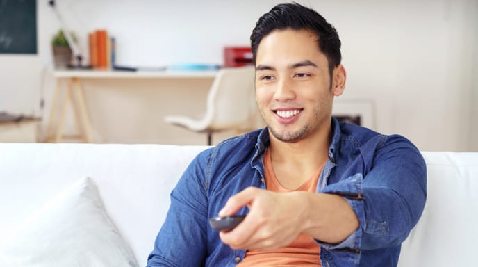 A man smiles while sitting on a couch, holding a TV remote.