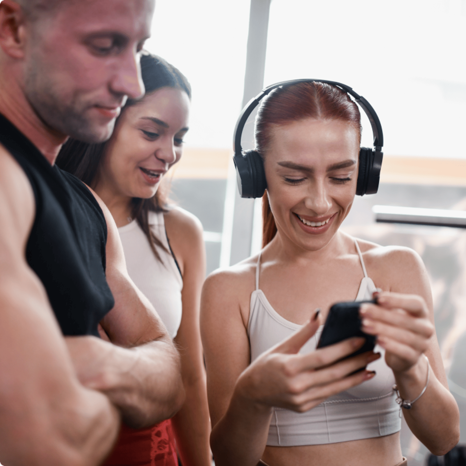 Eine Gruppe von drei Personen im Fitnessstudio, wobei eine lächelnde Frau mit Kopfhörern den anderen etwas auf ihrem Smartphone zeigt.