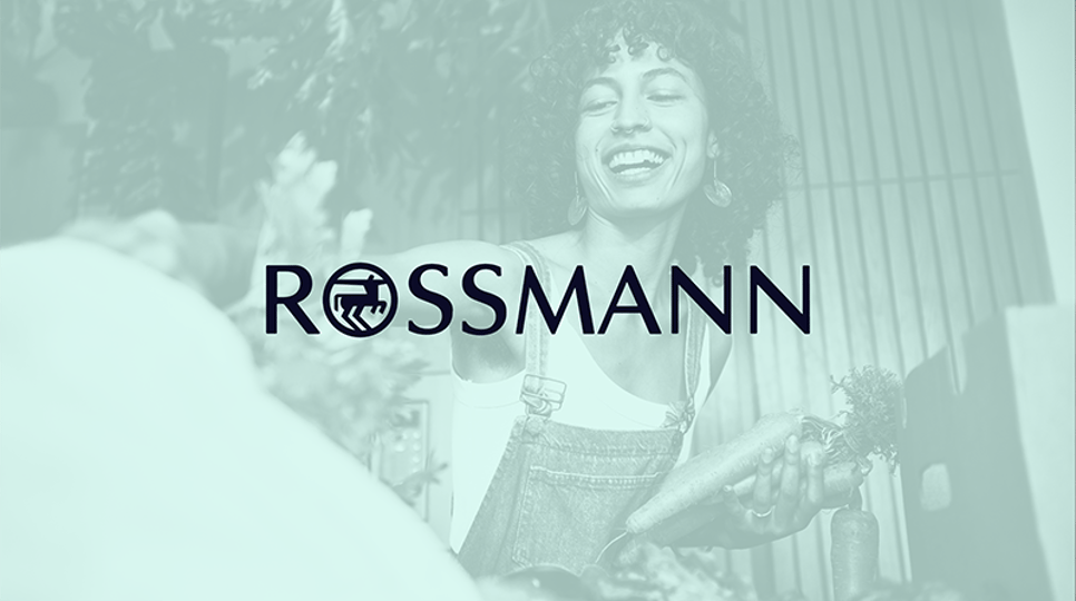 Eine Frau in Latzhose, sie lacht und packt Gemüse aus. Oben ist ein Rossmann-Logo platziert